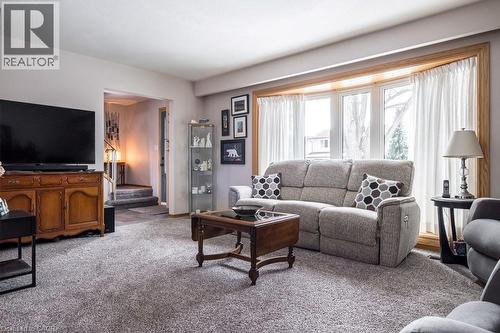 Living area featuring a bay window with wood trim, gray carpeting, neutral wall colors, and a partial view of a staircase - 19 Chateau Court, Hamilton, ON - Indoor Photo Showing Living Room