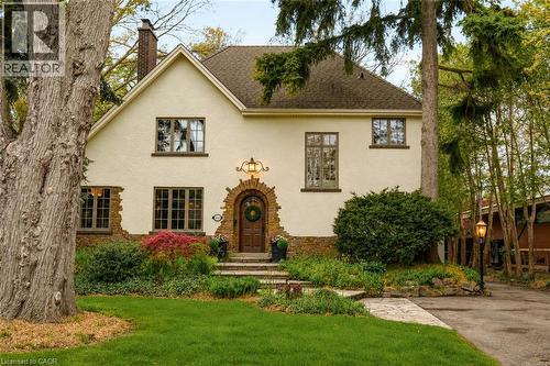 Stucco exterior residence featuring an arched wood-panel entry door, a stone pathway, a prominent chimney, and mature trees with established landscaping - 3081 Balmoral Avenue, Burlington, ON - Outdoor