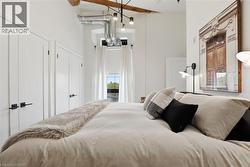Bedroom featuring two white paneled doors with matte black hardware, exposed ductwork, a large window with white curtains, and a prominent wooden beam - 