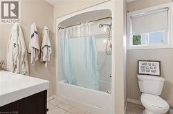 Bathroom featuring a tub/shower combination, a dark wood-finish vanity with an integrated white sink, a window with a roller blind, and light-toned floor tiles - 