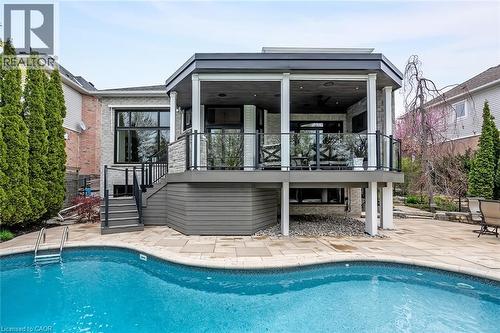 Curved swimming pool with stone paver surround, complemented by an elevated covered deck featuring glass railings and white support columns - 4285 Couples Crescent, Burlington, ON - Outdoor With In Ground Pool With Deck Patio Veranda