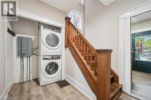 Wood-finish flooring with a stackable washer and dryer set, featuring an electrical panel, and a wooden staircase with a balustrade - 15 Gordon Avenue, Kitchener, ON - Indoor Photo Showing Laundry Room