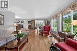 Spacious living area featuring polished wood-finish flooring, a wall of large windows, crisp white crown molding, light gray wall paint, and French doors opening to the sunroom - 