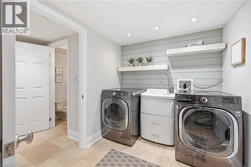 Laundry area featuring light-toned flooring, two front-loading laundry units, a utility sink, and wall-mounted shelving above a horizontally paneled accent wall - 3500 Regal Road, Burlington, ON - Indoor