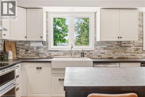 Kitchen featuring white shaker-style cabinetry, a farmhouse sink, stone tile backsplash, dark countertops, and stainless steel appliances - 2207 Jester Court, Burlington, ON - Indoor Photo Showing Kitchen With Double Sink