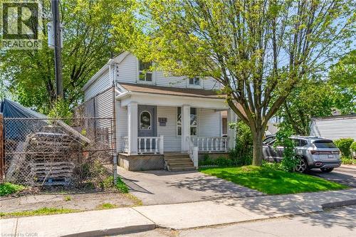 White siding residence featuring a covered front porch with white railing, a mature front yard tree, a paved driveway, and a chain-link fence - 168 Britannia Avenue, Hamilton, ON - Outdoor With Deck Patio Veranda