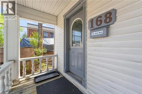 Front entry featuring a gray paneled door with an arched glass insert, white horizontal siding, and a covered porch with white turned balusters - 168 Britannia Avenue, Hamilton, ON - Outdoor With Exterior