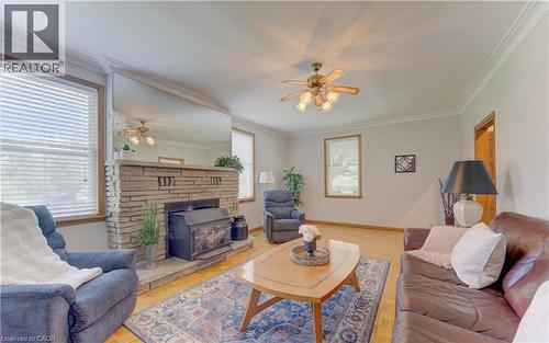 Living area featuring a stone-faced fireplace with a wood-burning insert, wood-finish flooring, a ceiling fan with light fixture, white crown molding, and two windows with blinds - 1463 Kossuth Road, Cambridge, ON - Indoor Photo Showing Living Room With Fireplace