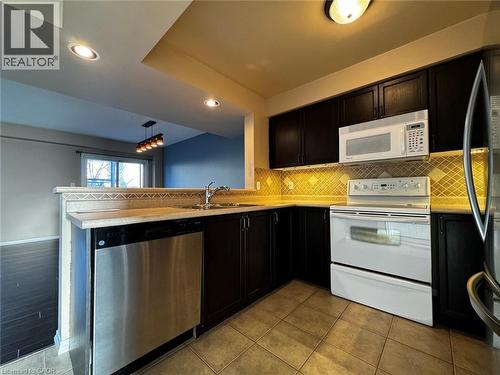 Kitchen featuring dark cabinetry, stainless steel dishwasher, white range with overhead microwave, tan tile flooring, and decorative tile backsplash - 37 Northcliffe Crescent, Cambridge, ON - Indoor Photo Showing Kitchen With Double Sink