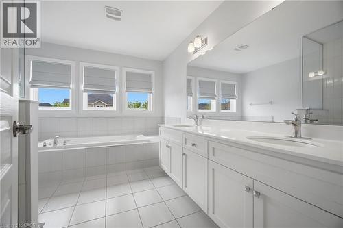 Bathroom featuring a double vanity with white cabinetry and integrated sinks, a built-in bathtub with tiled surround, three windows with blinds, and white square floor tiles - 58 Doon Creek Street, Kitchener, ON - Indoor Photo Showing Bathroom