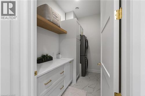 Dedicated laundry area featuring a stacked washer and dryer, white cabinetry with gold-tone hardware, a light-toned countertop, a floating wood shelf, and marble-look floor tiles - 2 Arbourvale Common Unit# 209, St. Catharines, ON - Indoor