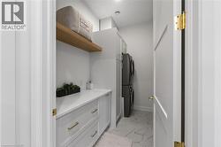 Dedicated laundry area featuring a stacked washer and dryer, white cabinetry with gold-tone hardware, a light-toned countertop, a floating wood shelf, and marble-look floor tiles - 