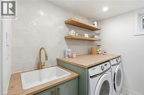 Dedicated laundry area featuring a built-in sink with a gooseneck faucet, wood-finish countertops, soft green cabinetry with gold hardware, two open shelving units, and a window for natural light - 345 Rankin Drive, Burlington, ON - Indoor Photo Showing Laundry Room