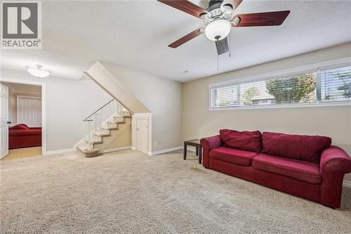 Spacious carpeted room featuring a ceiling fan with integrated lighting, multiple transom windows with blinds, a staircase with white balusters, and a recessed under-stair storage door - 47 Oakhurst Crescent, Kitchener, ON - Indoor