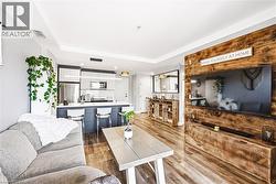 Open-concept living space featuring wood-finish flooring, a contemporary kitchen island with seating, stainless steel appliances, a linear pendant light fixture, and a distinctive wood-paneled accent - 