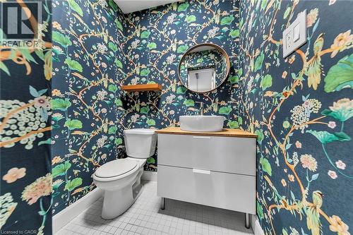 Powder room featuring botanical wallpaper, a white vessel sink, a light wood-finish countertop, a round frameless mirror, and white square floor tiles - 501 Ferguson Avenue N, Hamilton, ON - Indoor Photo Showing Bathroom