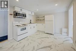 Modern kitchen featuring white cabinetry with dark hardware, stainless steel microwave, white electric range, white refrigerator, and marble-look tile flooring - 