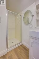 Compact bathroom featuring a glass-enclosed shower with a white base, wood-finish flooring, a contemporary white vanity with integrated sink, and an oval wall-mounted mirror - 