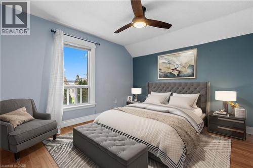 Wood-finish flooring with a multi-tone area rug, featuring a large window with white trim, a ceiling fan with integrated lighting, and a prominent accent wall (Virtually Staged) - 204 Beach Boulevard, Hamilton, ON - Indoor Photo Showing Bedroom