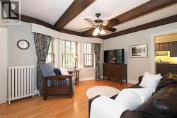 Living area featuring wood-finish flooring, exposed ceiling beams, a bay window with natural light, a ceiling fan, and a radiator - 