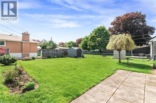 Expansive grass lawn featuring a concrete patio, perimeter chain-link fencing, mature deciduous trees, and two storage sheds - 435 East 36Th Street, Hamilton, ON - Outdoor