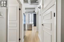 Hallway with light wood-finish flooring, white paneled doors, exposed industrial-style ceiling, stainless steel dishwasher, and built-in wall sconce - 