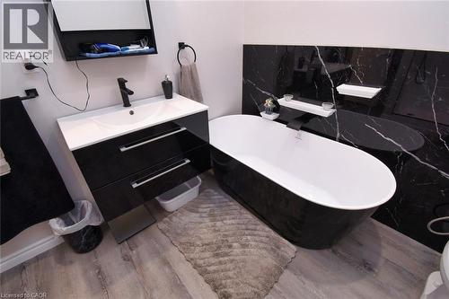 Bathroom featuring a freestanding black and white soaking tub, a wall-mounted vanity with integrated sink and black cabinetry, black accent wall with white veining, wood-finish flooring, and matte bla - 5 Ralph Street, Turkey Point, ON - Indoor Photo Showing Bathroom