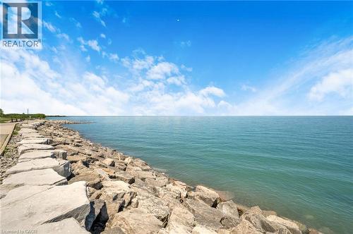 Expansive waterfront featuring a natural stone breakwater, tranquil teal water, clear blue sky, and a paved shoreline path - 10 Esplanade Lane Unit# 310, Grimsby, ON - Outdoor With Body Of Water With View