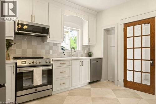 Modern kitchen featuring white shaker cabinetry, stainless steel appliances, light-toned tiled flooring, a subway tile backsplash, and a wood-frame glass panel door - 106 Grosvenor Avenue N, Hamilton, ON - Indoor Photo Showing Kitchen