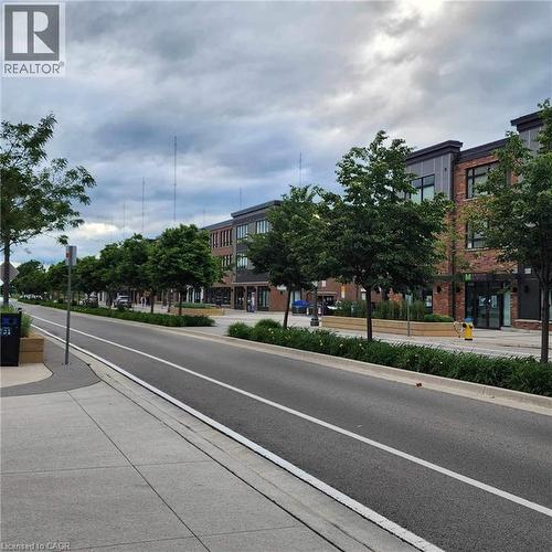 Urban streetscape featuring modern brick and panel-sided buildings, a paved roadway with a designated lane, a concrete sidewalk, and landscaped planters with mature trees and shrubbery - 385 Winston Road Unit# 709, Grimsby, ON - Outdoor