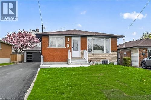 Brick and stone facade bungalow featuring a paved driveway, attached garage, front lawn, and mature flowering tree - 74 Victor Boulevard, Hamilton, ON - Outdoor
