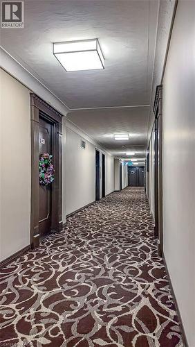 Long hallway featuring patterned carpet, multiple ceiling-mounted light fixtures, dark wood door frames, and light-colored walls - 40 Harrisford Street Unit# 802, Hamilton, ON 