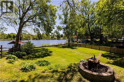 Expansive waterfront backyard featuring mature trees, a manicured lawn, a block-built fire pit, and a wooden fence - 100 Mechanic Street W, Waterford, ON - Outdoor With Body Of Water