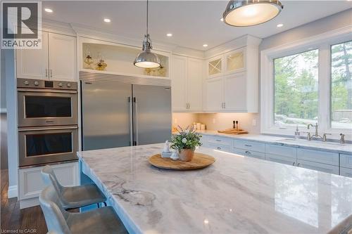 Kitchen featuring a spacious island with a natural stone countertop, white shaker cabinetry, stainless steel double ovens, a stainless steel refrigerator, and ample natural light from large windows - 1521 Emily Lane, Severn, ON - Indoor Photo Showing Kitchen With Upgraded Kitchen