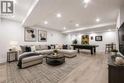 Spacious lower-level entertainment space featuring light wood-finish flooring, recessed ceiling lights, and white baseboards - 