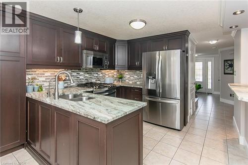 Kitchen featuring dark wood-finish cabinetry, stainless steel appliances, mosaic tile backsplash, and a central island with a double basin sink and a stone countertop - 529 Chesapeake Crescent, Waterloo, ON - Indoor Photo Showing Kitchen With Double Sink With Upgraded Kitchen