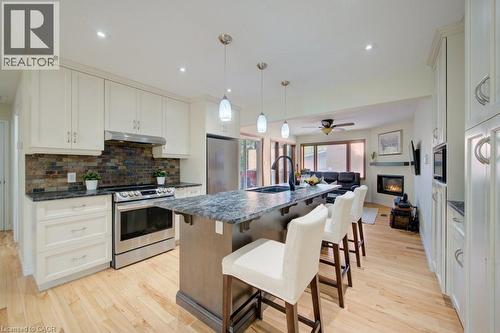 Contemporary kitchen featuring a large central island with seating, stainless steel appliances, light wood-finish flooring, white cabinetry, and a stacked stone backsplash - 60 Mayfield Avenue, Waterloo, ON 