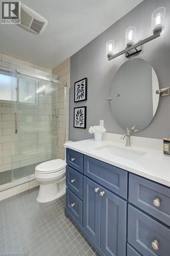 Bathroom featuring a navy blue vanity with a white countertop and integrated sink, a glass-enclosed shower with subway tile surround, an oval mirror, and a three-light vanity fixture - 60 Mayfield Avenue, Waterloo, ON 