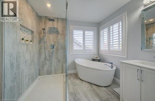 Bathroom featuring a glass-enclosed shower with tile surround, a freestanding soaking tub, two windows with white plantation shutters, a single vanity with white cabinetry, and wood-finish tile floori - 7 Housler Lane, Cambridge, ON 