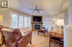 Living area featuring wood-finish flooring, a corner fireplace with stone surround and wood mantel, white plantation shutters, and a ceiling fan - 
