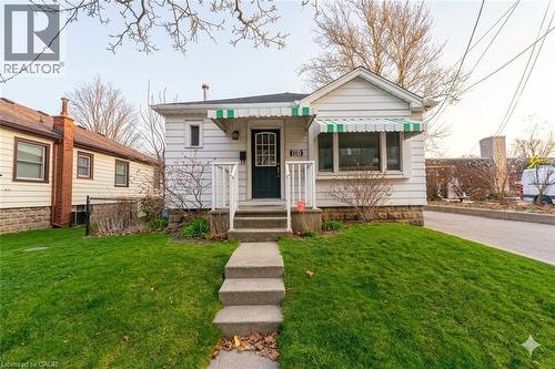 White siding exterior featuring a dark entry door, front porch with white railing, and dual green and white striped awnings - 501 Upper Wellington Street, Hamilton, ON 