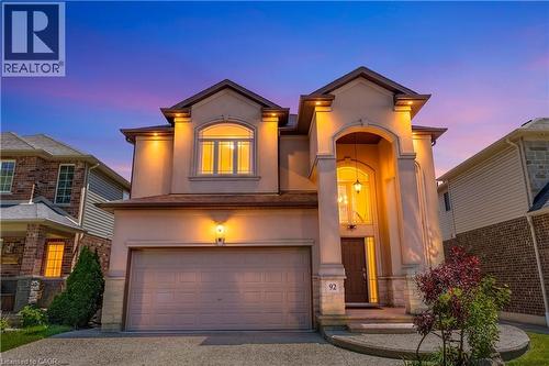 Stucco exterior with stone accents, featuring an arched entryway with a hanging pendant light, an arched second-story window, a two-car garage, and exterior uplighting - 92 Desoto Drive, Hamilton, ON - Outdoor