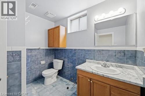 Bathroom featuring blue tile wainscoting and flooring, a window, a single vanity with a white countertop and wood-finish cabinetry, and a wall-mounted mirror with an overhead light fixture - 957 Stone Church Road E, Hamilton, ON - Indoor Photo Showing Bathroom