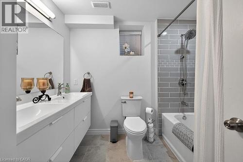Bathroom featuring a dual-sink vanity with under-sink storage, a subway tile shower surround with an accent band, a white bathtub, and light-toned floor tiling - 109 Ray Street N, Hamilton, ON - Indoor Photo Showing Bathroom