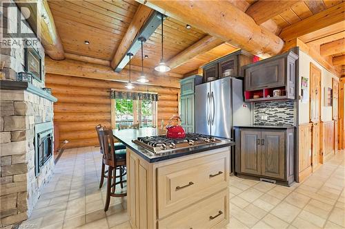 Rustic kitchen and living area featuring a central island with a built-in gas range, a stone-clad fireplace, log wall construction, exposed log beams, and a tile backsplash - 169 James Street, Atwood, ON - Indoor Photo Showing Kitchen