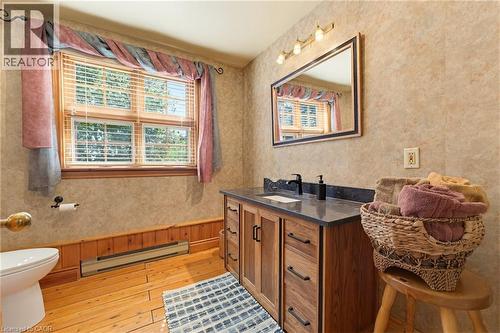 Bathroom featuring a wood-finish vanity with a dark countertop and integrated sink, a large framed mirror with overhead lighting, a window with horizontal blinds, wood-finish flooring, and wood paneli - 169 James Street, Atwood, ON - Indoor Photo Showing Bathroom