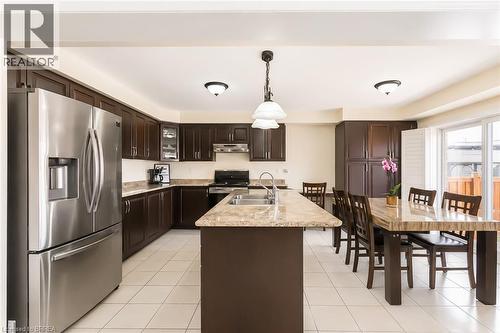 Open-concept kitchen featuring dark wood cabinetry, stainless steel appliances, light-toned countertop surfaces, a kitchen island with an integrated sink, and white tile flooring - 26 Pollard Street, Brantford, ON - Indoor