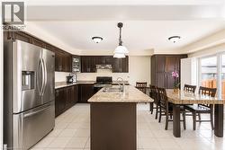 Open-concept kitchen featuring dark wood cabinetry, stainless steel appliances, light-toned countertop surfaces, a kitchen island with an integrated sink, and white tile flooring - 