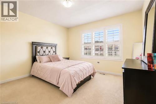 Carpeted room featuring a ceiling-mounted light fixture, light yellow walls, white baseboards, and a triple window with white plantation shutters - 26 Pollard Street, Brantford, ON - Indoor Photo Showing Bedroom