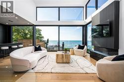 Spacious living area featuring expansive floor-to-ceiling windows, high ceilings, wood-finish flooring, a contemporary fireplace with a dark stone surround, and a built-in bar area - 